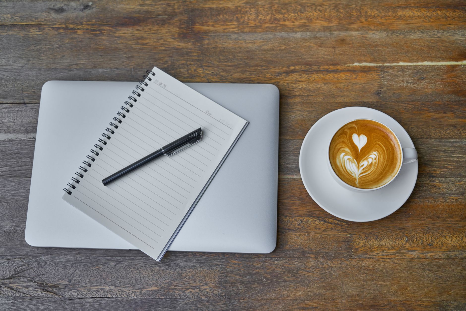 A coffee cup, notebook, pen and laptop on a rustic wooden table