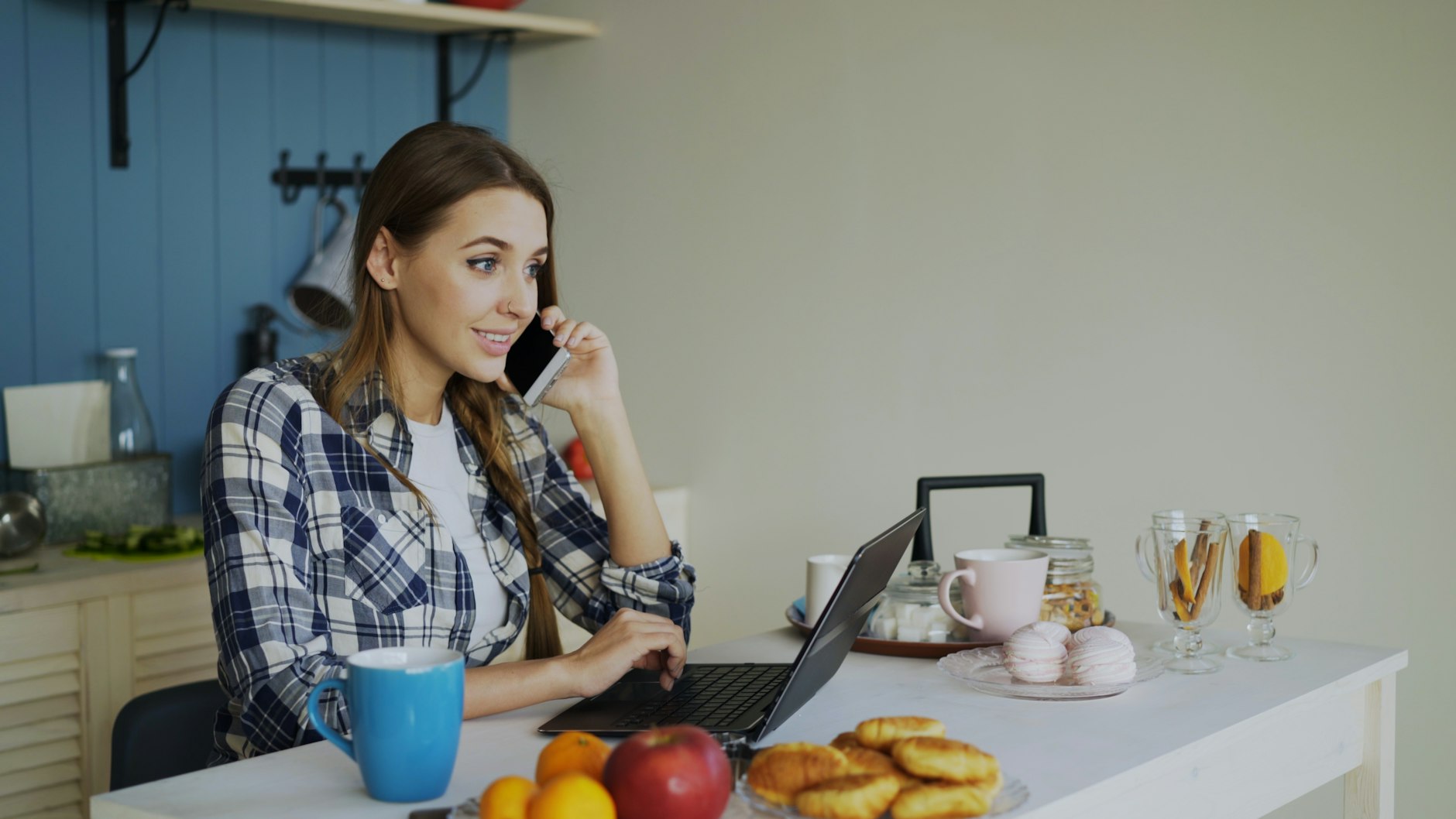 A woman on the phone while working on a laptop at her kitchen table